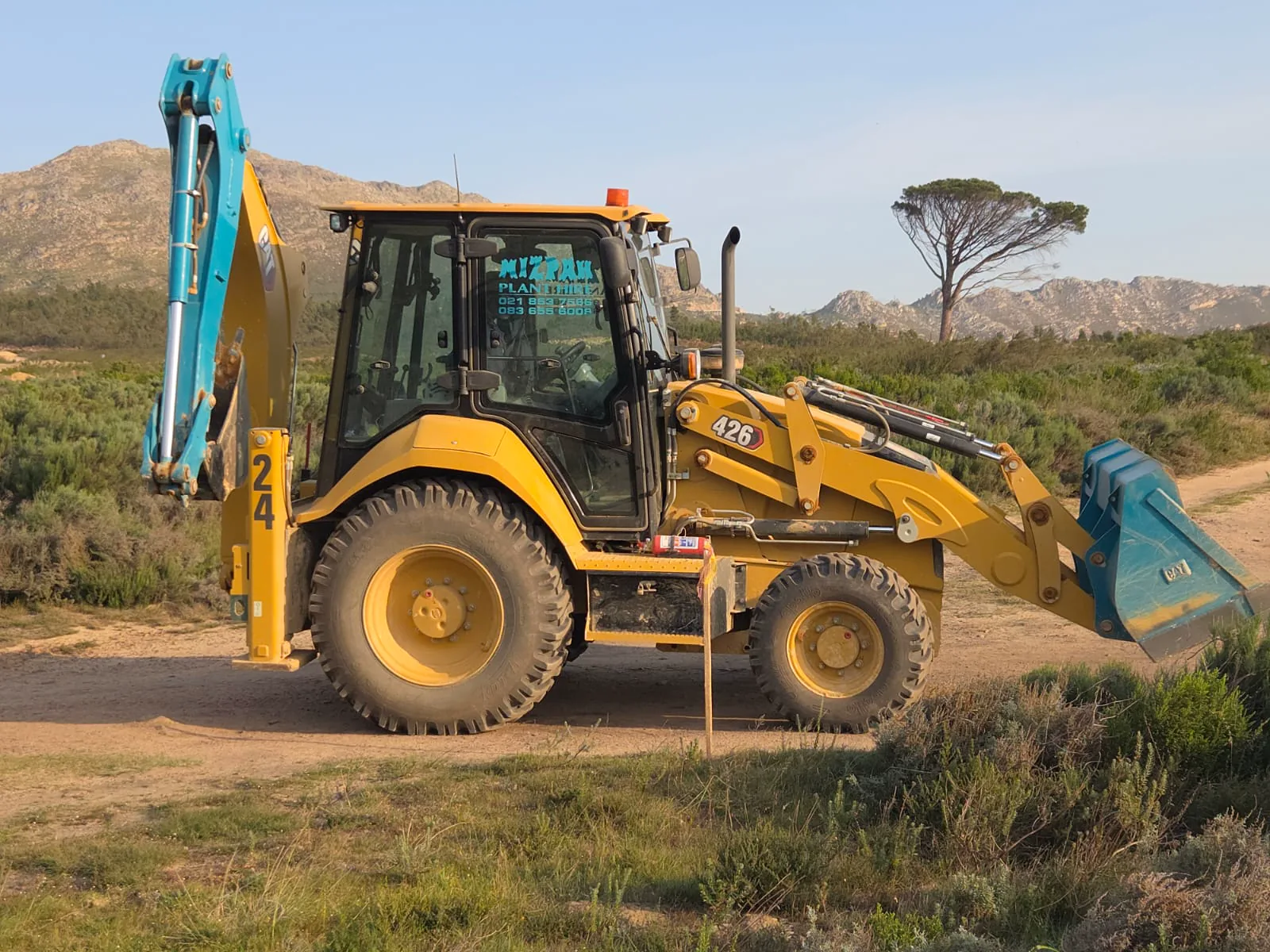 Side profile of a Mizpah Plant Hire TLB with mountains and a tree in the background
